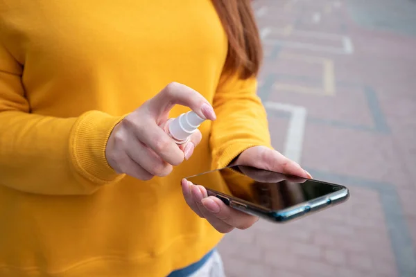 Sarı kazaklı genç bir kız telefonunu sprey dezenfektanla dezenfekte ediyor.
