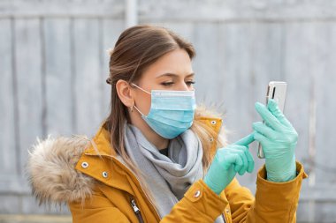 Koruyucu maskeli ve eldivenli genç bir kadının portresi elinde akıllı telefon, koronavirüs salgını alarmı.