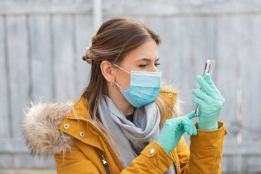 Koruyucu maskeli ve eldivenli genç bir kadının portresi elinde akıllı telefon, koronavirüs salgını alarmı.