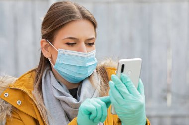 Koruyucu maskeli ve eldivenli genç bir kadının portresi elinde akıllı telefon, koronavirüs salgını alarmı.