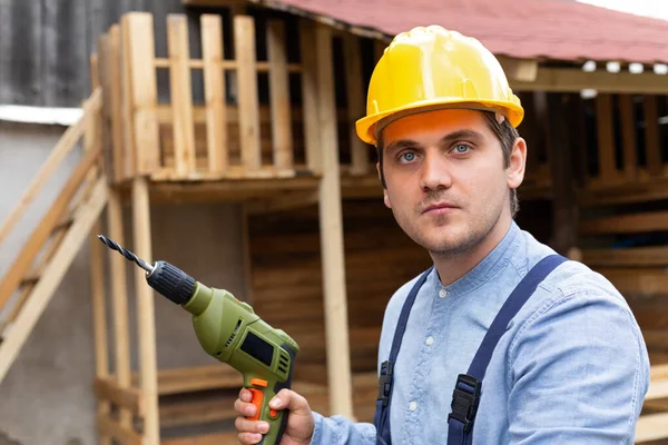 Young handsome carpenter with yellow hard hat and work equipment is holding an electric drill on wooden background