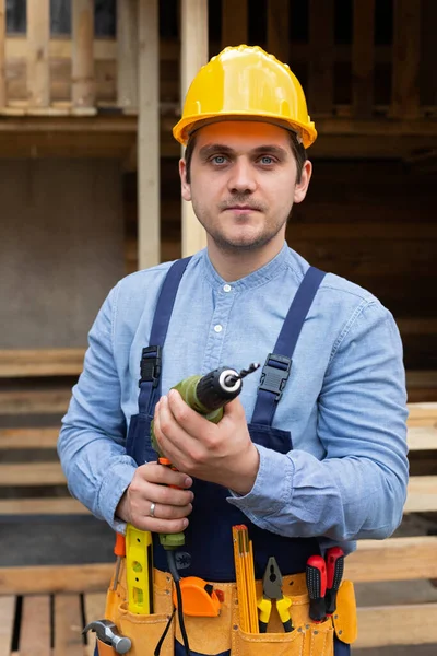 Young handsome carpenter with yellow hard hat and work equipment is holding an electric drill on wooden background