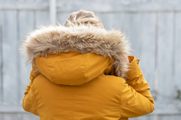 Back view of woman wearing yelllow furry parka jacket in front of grey wooden background