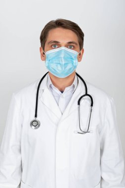 Cerrahi maske takan yakışıklı genç doktor beyaz arka planda kameraya bakıyor.