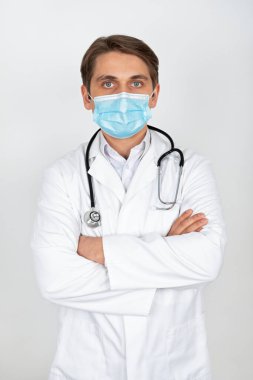 Cerrahi maske takan yakışıklı genç doktor beyaz arka planda kameraya bakıyor.