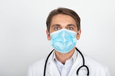 Cerrahi maske takan yakışıklı genç doktor beyaz arka planda kameraya bakıyor.