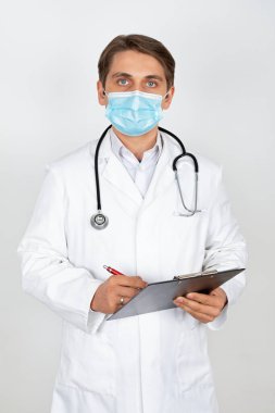 Cerrahi maske takan yakışıklı genç doktor beyaz arka planda kameraya bakıyor.