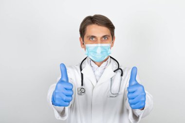 Cerrahi maske ve eldiven takan yakışıklı genç doktor beyaz arka planda baş parmak gösteriyor.