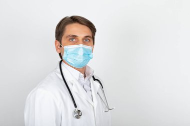 Cerrahi maske takan yakışıklı genç doktor beyaz arka planda kameraya bakıyor.
