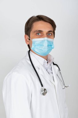 Cerrahi maske takan yakışıklı genç doktor beyaz arka planda kameraya bakıyor.