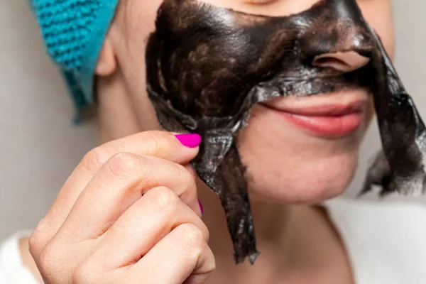 Close up picture of woman removing black charcoal peel off face mask from skin - pore cleansing treatment at home