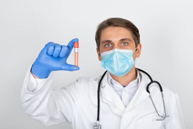 Cerrahi maskeli ve eldivenli genç bir erkek doktor beyaz arka planda kan örneği tutuyor.