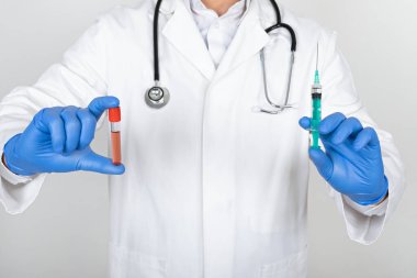 Ameliyat maskesi ve eldivenleri olan genç bir erkek doktor elinde kan örneği ve beyaz arka planda iğne tutuyor.