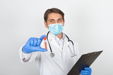 Cerrahi maskeli ve eldivenli genç bir erkek doktor beyaz arka planda kan örneği tutuyor.