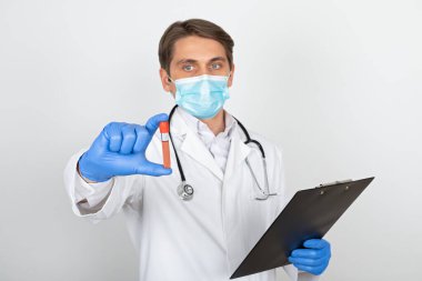 Cerrahi maskeli ve eldivenli genç bir erkek doktor beyaz arka planda kan örneği tutuyor.