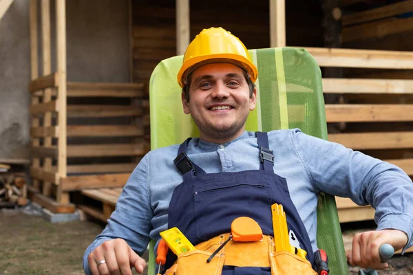 Picture of happy young carpenter with uniform and yellow helmet resting after hard work outdoor