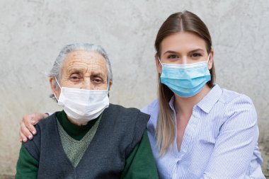 Arkadaş canlısı bakıcı portresi. Yaşlı hasta kadınla poz veriyor. Covid-19 salgını yüzünden cerrahi maske takıyor.