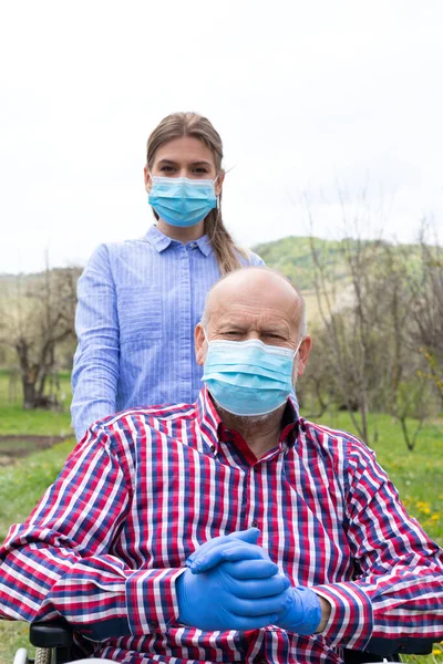 Elderly disabled man with mask sitting in wheelchair, assisted by young female caregiver outdoors