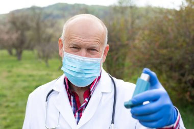 Cerrahi maskeli yaşlı erkek doktor dışarıda astım spreyi tutuyor.
