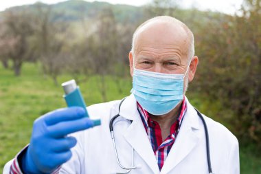 Cerrahi maskeli yaşlı erkek doktor dışarıda astım spreyi tutuyor.