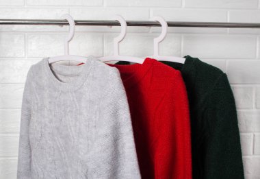 Elbiseler askıda asılı. Kırmızı, yeşil ve melanj renkli kazaklar. Moda ve tarz, vitrin, mağaza, konsept. Beyaz tuğla duvar, konfor ve temizlik, tasarım, kızlık zarı, yumuşak ve örgü. Dolap.