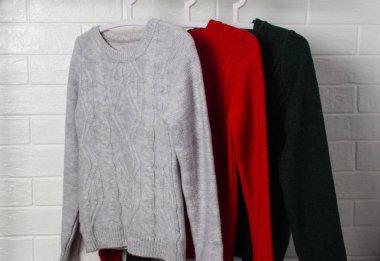 Elbiseler askıda asılı. Kırmızı, yeşil ve melanj renkli kazaklar. Moda ve tarz, vitrin, mağaza, konsept. Beyaz tuğla duvar, konfor ve temizlik, tasarım, kızlık zarı, yumuşak ve örgü. Dolap.