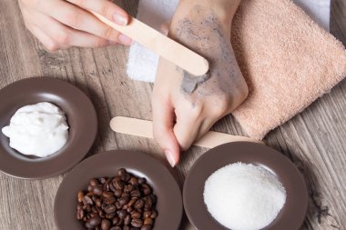 Kadının biri eline ev yapımı bir spatula sürmüş. SPA, güzellik. Fırça, kahve, şeker, krema yapmak için gerekli malzemeler. Cilt bakımı. Nemlendirici, deri yüzücü, soyucu. Ev Kozmetikleri, Eko