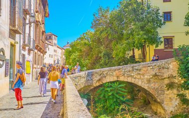 GRANADA, SPAIN - SEPTEMBER 25, 2019: People walk arched Puente Espinosa bridge over Darro river and stone walkway of Carrera del Darro street of Albaicin district, on September 25 in Granada