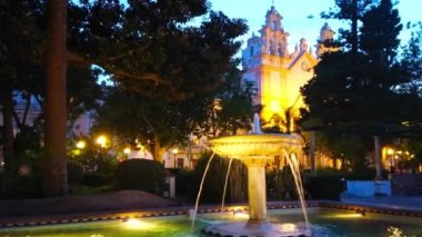 Alameda Marques de Comillas bahçesinde klasik taş çeşme manzaralı ve arka planda Carmen Kilisesi 'ni aydınlatan gece, Cadiz, İspanya