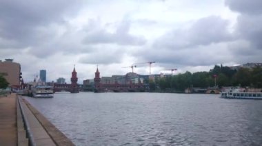 BERLİN, GERMANY - 3 Ekim 2019: Spree nehrinin yanında yürüyün ve 3 Ekim 'de Berlin' de kırmızı kuleler, eğlence tekneleri ve bulutlu gökyüzü ile tarihi Oberbampagne ucke (Oberbaum Köprüsü) 'ü izleyin