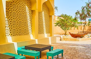 The shady street with wooden benches and tables, Arabic screen panels with fine Islamic patterns and vintage fishing boat amid the flower bed, Al Bastakiya (Al Fahidi) district, Dubai, UAE clipart