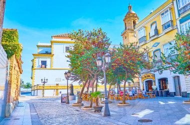 CADIZ, SPAIN - 23 Eylül 2019: 23 Eylül 'de Cadiz' de arka planda gür ağaçları, eski yapıları, açık kafesi ve Santo Domingo kilisesi olan gölgeli Calle Plocia caddesi