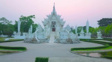 Büyüleyici Beyaz Tapınak (Wat Rong Khun) üzerindeki mor alacakaranlık, karmaşık pervazlar, ayna mozaiği, heykeller ile süslenmiş ve göl ve çalı bahçesi ile çevrili, Chiang Rai, Tayland