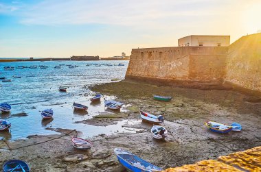 Santa Catalina Kalesi 'nin ortaçağ taş surları üzerinde parlak günbatımı gökyüzü ve eski botlarla sığ La Caleta limanı, Cadiz, İspanya