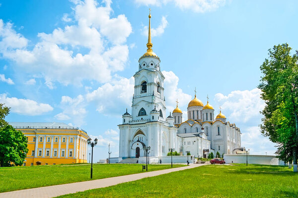 The splendid Dormition (Assumption) Cathedral with carved tall bell tower, white walls and golden domes amid the greenery of Pushkin Park, Vladimir, Russia