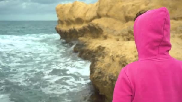 Una giovane donna è in piedi di fronte a una scogliera nel mare. Il mare tra le rocce. Una donna sta con la schiena con una felpa rosa. Tempesta sul Mar Mediterraneo. — Video Stock