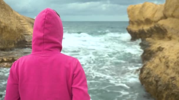 Egy fiatal nő áll egy szikla előtt a tengerben. A tenger a sziklák között. Egy nő áll a háta mögött rózsaszín kapucnis pulcsiban. Vihar a Földközi-tengeren. — Stock videók