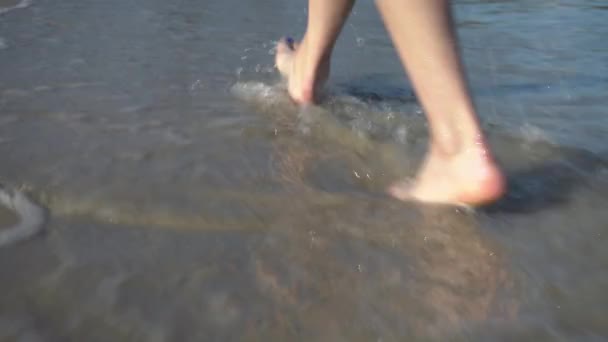 Una mujer camina sobre la arena en la playa. Las piernas cierran. Las olas llegan a la orilla del mar — Vídeos de Stock