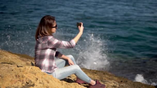 Una joven está sentada con un teléfono en las piedras frente al mar. Ella misma lleva gafas de sol. Salpicaduras de olas marinas. Movimiento lento . — Vídeos de Stock