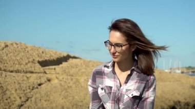 Genç bir kadın denizin arka planında taşların üzerinde oturuyor. Kız rüzgarda saçlarını sallıyor. Güneş ışıl ışıl parlıyor. Yavaş çekim