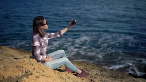 Uma jovem está sentada com um telefone nas pedras em frente ao mar. Uma rapariga com óculos de sol. Splashes de onda do mar — Vídeo de Stock