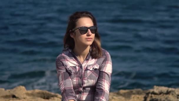 Une jeune femme est assise sur des pierres sur un fond de mer. Fille en lunettes de soleil par temps ensoleillé. Explore la nature environnante — Video