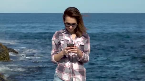 Una giovane donna sta con un telefono sullo sfondo del mare. Donna che scrive sullo smartphone durante le vacanze. Tempo soleggiato — Video Stock