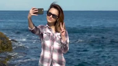 Genç bir kadın denizin arka planında akıllı bir telefondan selfie çekiyor. Kadın tatilde fotoğraf çekiyor. Güneşli havada mavi deniz