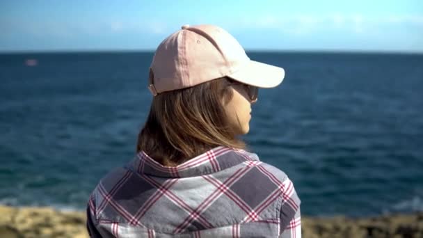 Eine junge Frau blickt aufs Meer. Das Mädchen mit der Mütze bewundert die Natur. Frau steht mit dem Rücken zur Kamera. — Stockvideo