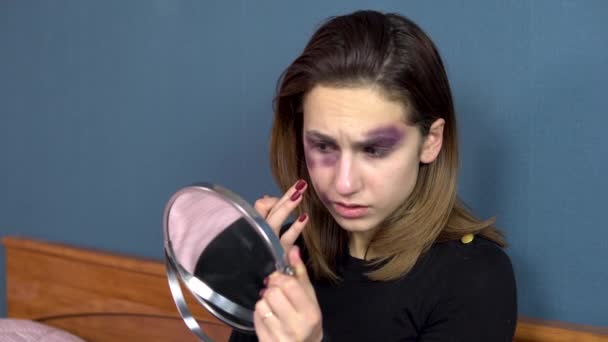 A young woman examines bruises on her face in the mirror. Quarrels in a young family. Domestic violence. The girl is sitting on the bed — Stock Video