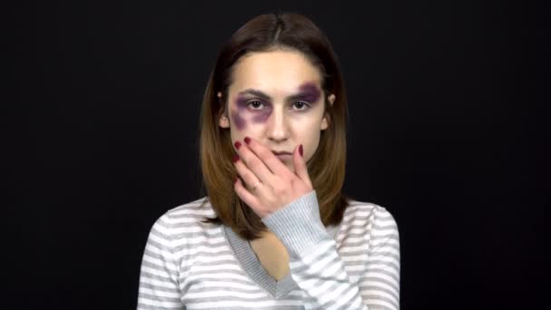 Eine junge Frau berührt blaue Flecken im Gesicht und blickt in die Kamera. Streit in einer jungen Familie. Häusliche Gewalt. auf schwarzem Hintergrund — Stockvideo