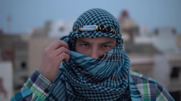 Νεαρός με αραβικό μαντήλι. Ένας άντρας κοιτάζει την κάμερα και ανοίγει ένα μαντήλι. Στην οροφή του σπιτιού με φόντο τα αραβικά σπίτια. — Αρχείο Βίντεο