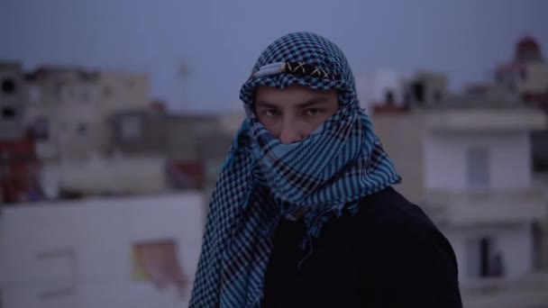 Młody człowiek w arabskiej chusteczce. Mężczyzna patrzy w kamerę i wychodzi. Na dachu domu na tle arabskich domów. — Wideo stockowe