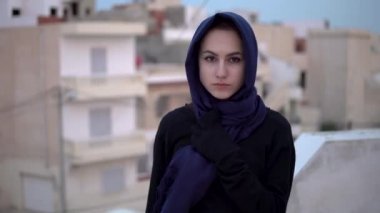 Arap şallı genç bir kadın. Bir kadın kameraya bakıyor. Evin çatısında, Arap evlerinin arka planına karşı..